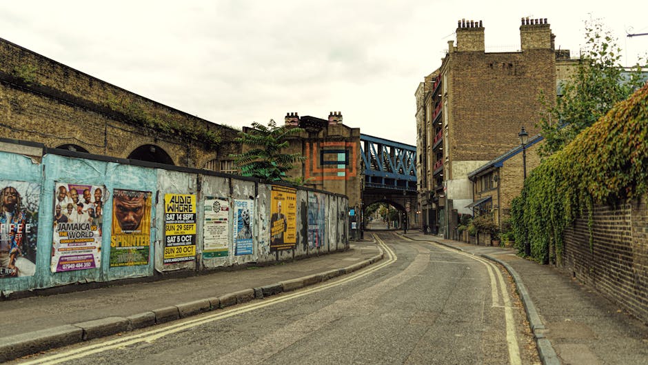 A narrow curving street in an urban area features a weathered blue metal fence on the left side, covered with various colorful posters advertising events. The road has a double yellow line along the c